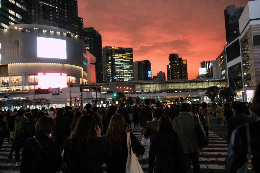 Pedestrians flooding a wide Tokyo intersection from every direction under bright advertising screens at night