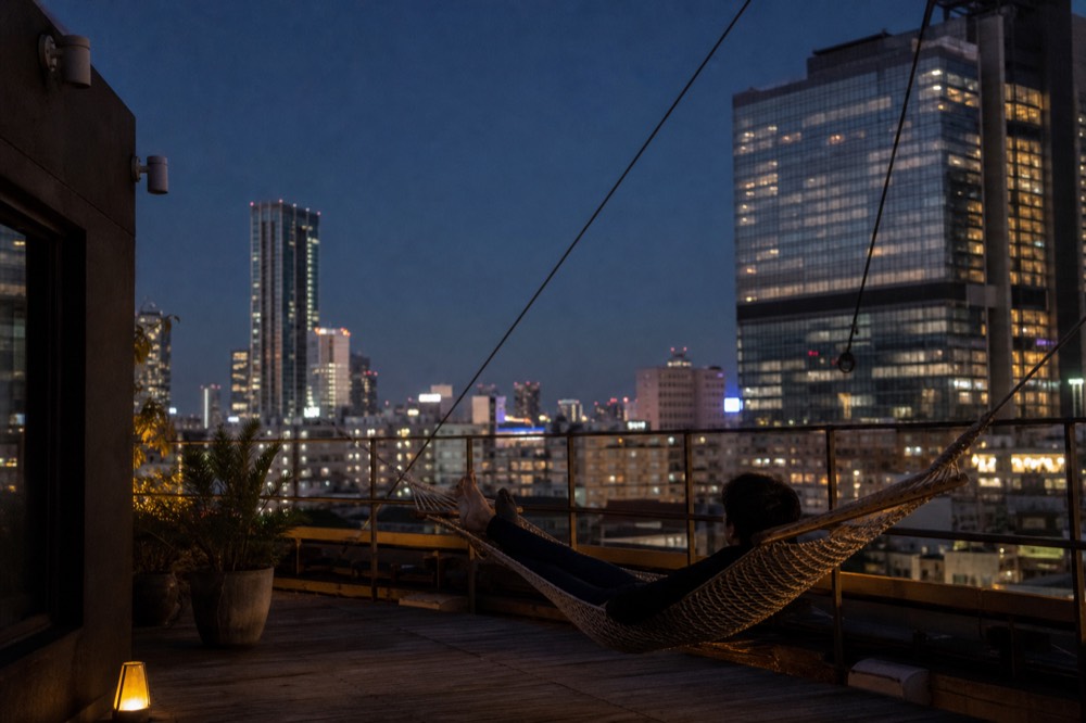 Person reclining in a suspended rope hammock on a high rooftop with open evening sky above and distant towers beyond