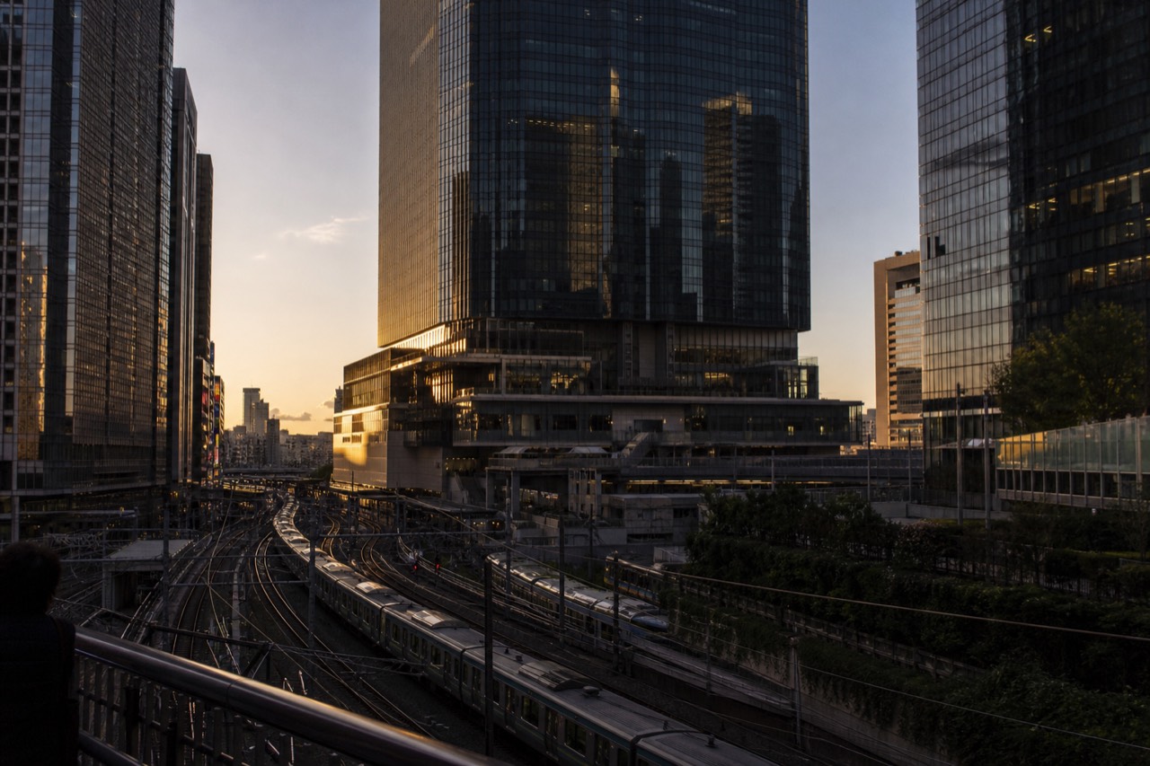 Tall dark glass tower rising directly above a busy Tokyo railway station at dusk, train lines curving away beneath it