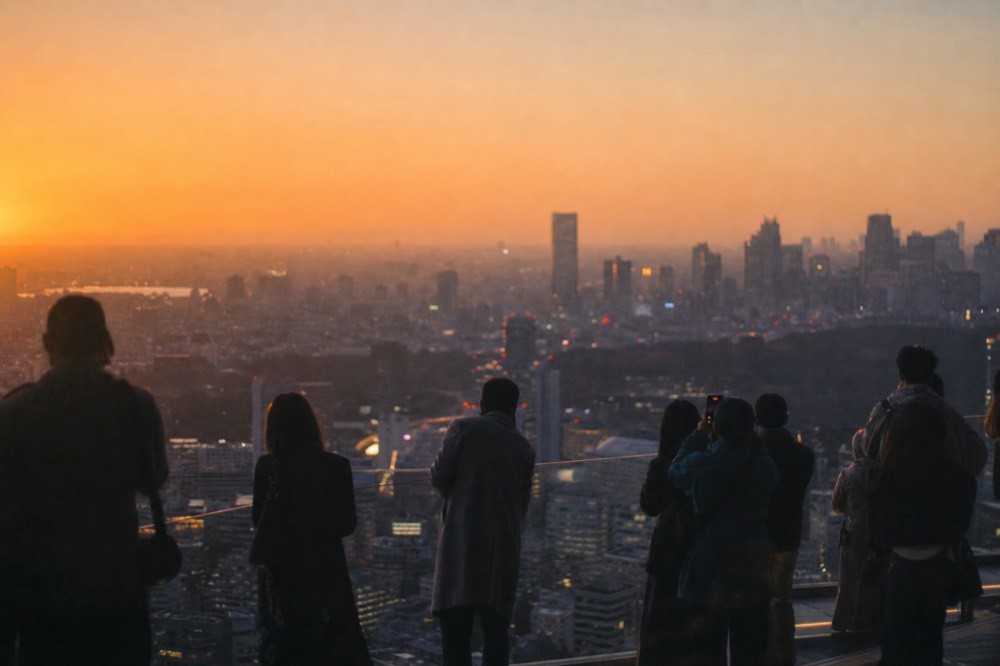 Crowd of silhouetted visitors along a glass rooftop parapet at sunset with an orange sky over a dense city skyline