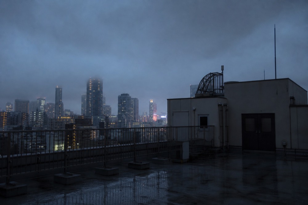 Empty wet rooftop deck under heavy grey cloud with closed access doors and city towers half lost in low mist