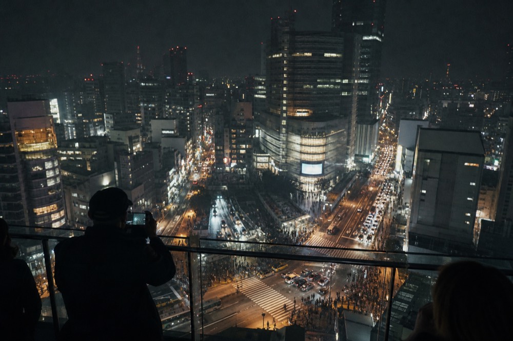 Night view from a high rooftop over a famous pedestrian scramble crossing, streets glowing with white headlights and coloured signage