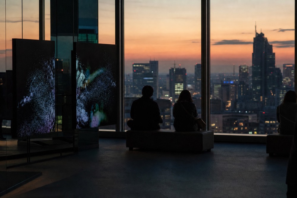 Indoor glass-walled observation gallery on a high floor at dusk with dark digital art panels and visitors seated facing the city