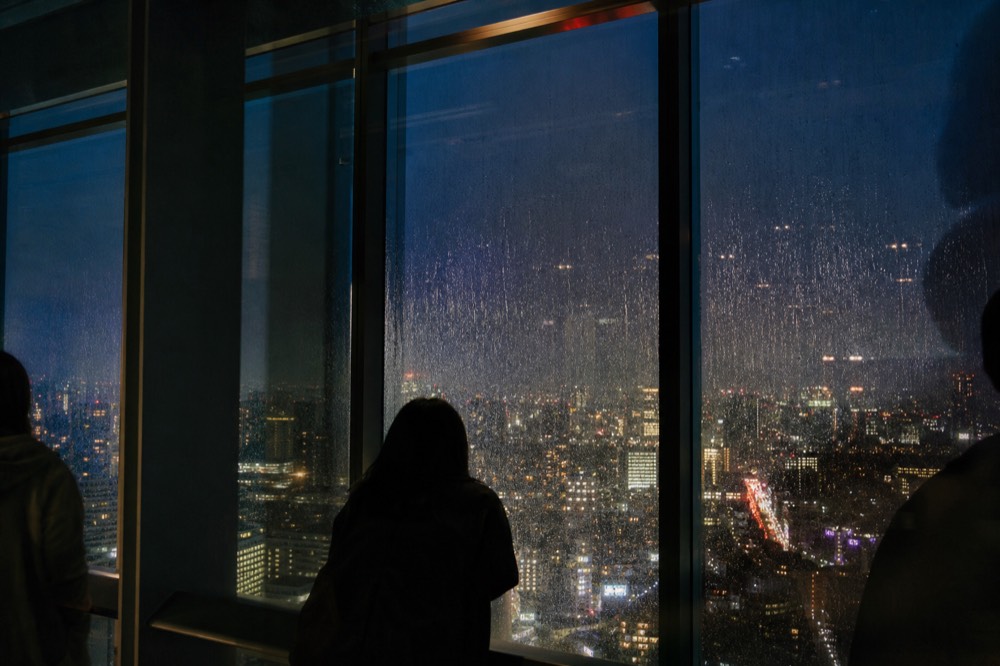 Rain streaking across the glass of a high indoor observation gallery with the city below reduced to blurred coloured light